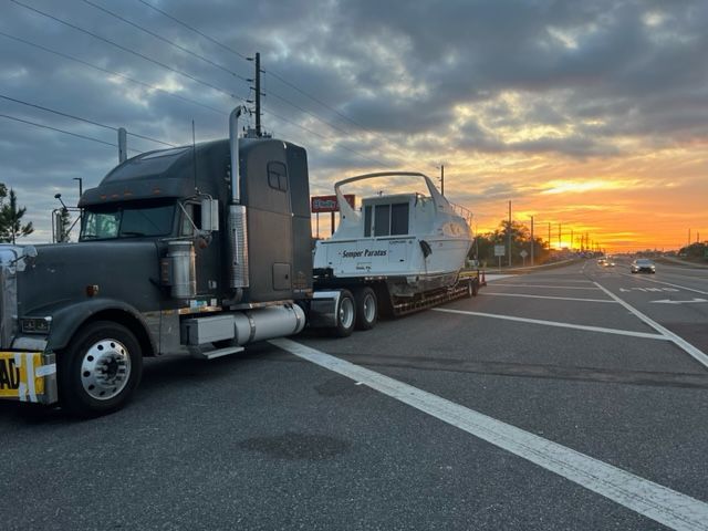 Semi-truck transporting a boat on a trailer across a road at sunset.