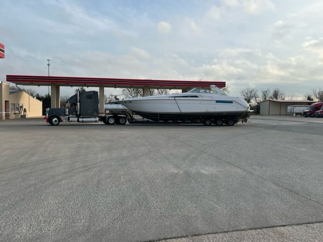 A semi-truck transports a large white boat on a trailer at a gas station.