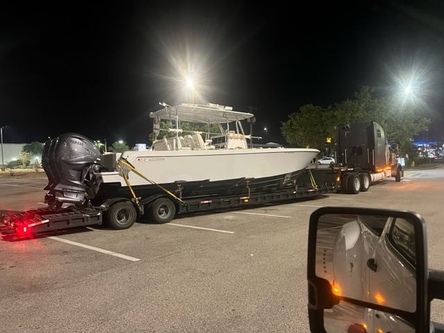 A large white boat on a trailer, being towed by a semi-truck in a parking lot at night.