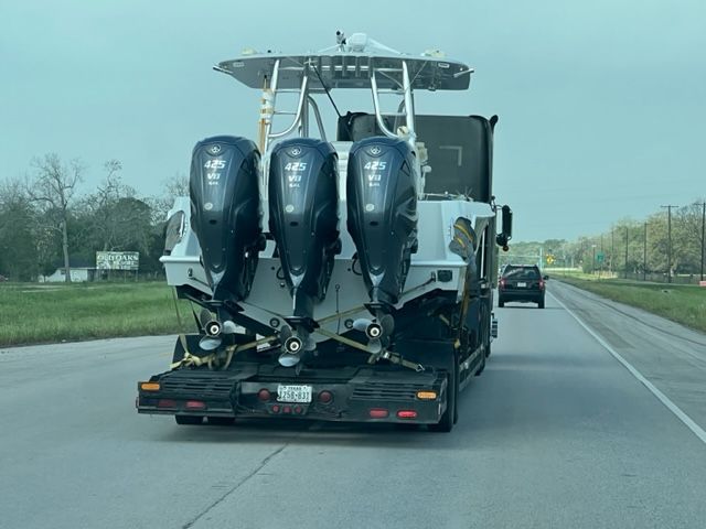 Boat on a trailer being transported down a highway. Three large outboard motors are visible.