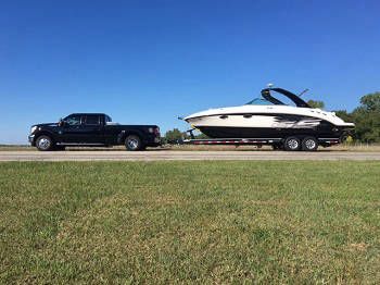 Black truck towing a white boat on a trailer across a green field under a blue sky.