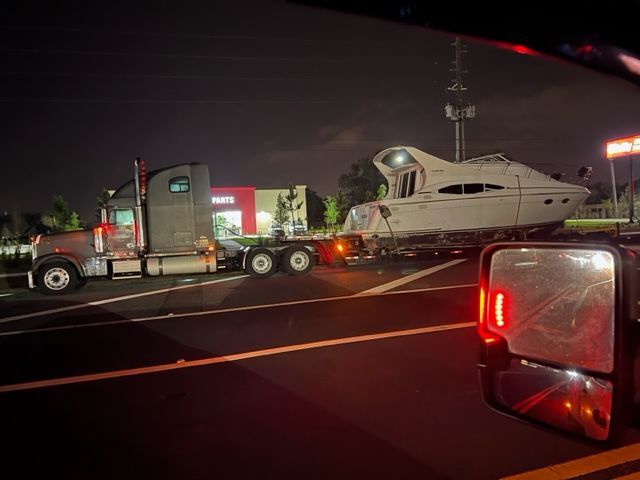 A semi-truck transports a large white yacht at night. The truck is next to a business with red signage.