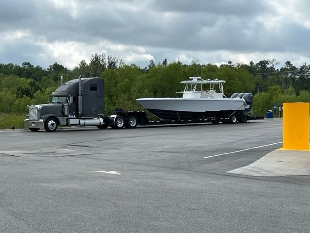 A semi-truck hauling a large gray boat on a trailer, parked near trees and a yellow pole.