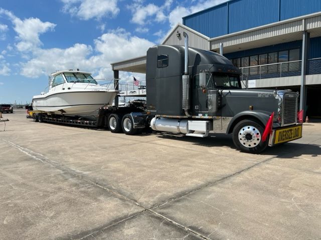 A large boat on a trailer is pulled by a gray semi-truck on a paved lot, under a blue sky.
