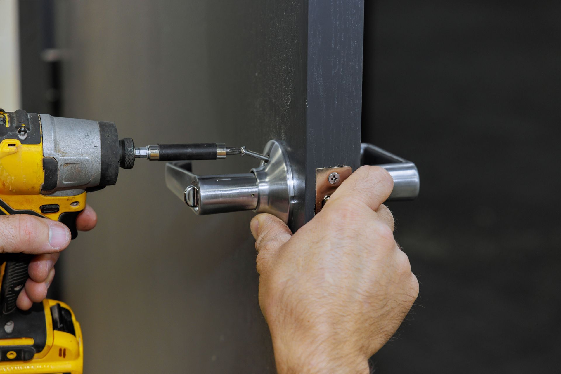 A person uses a yellow power drill to install a metal door handle on a dark gray door.