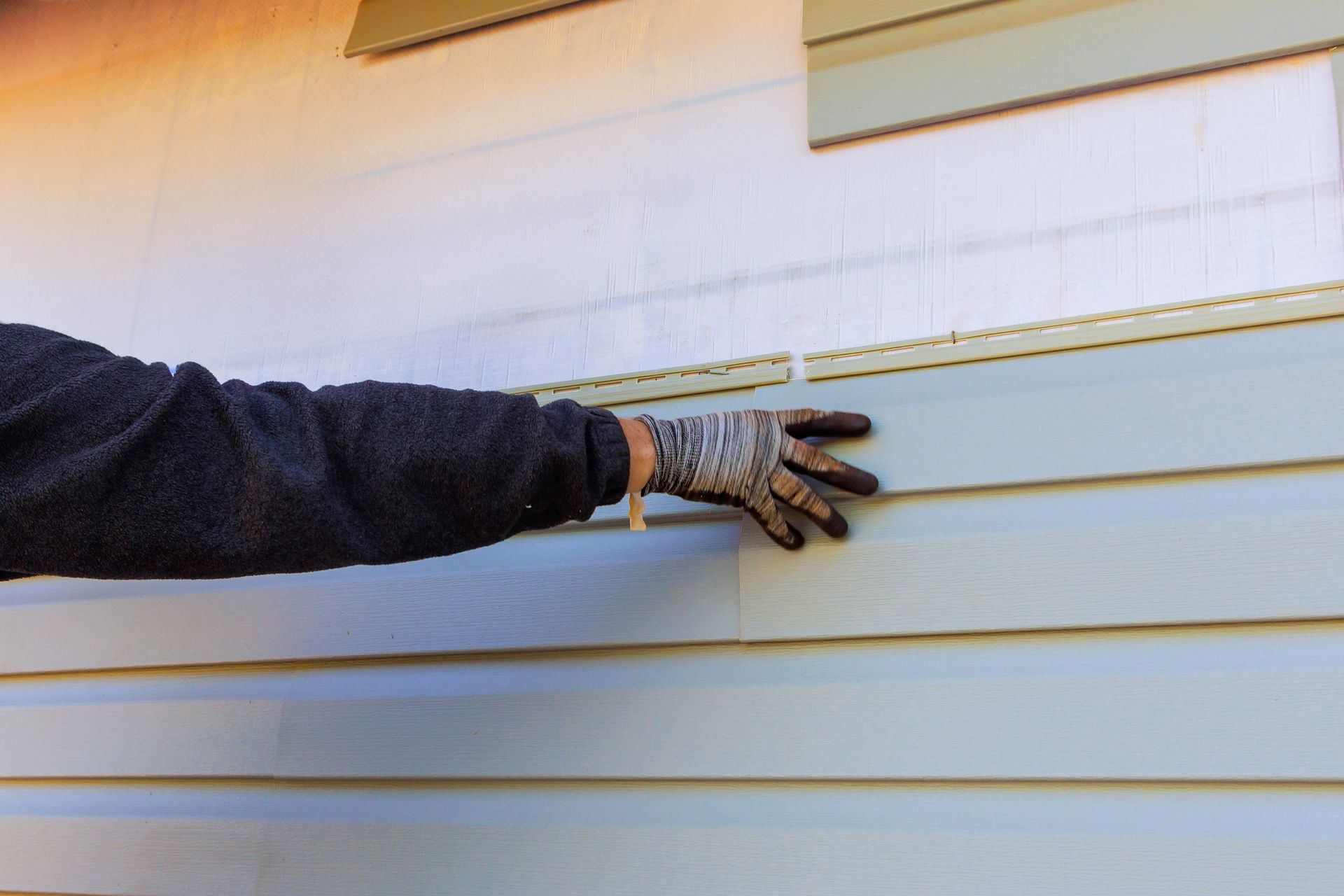 A person wearing a work glove presses their hand against a light blue horizontal siding panel installed on a house wall.