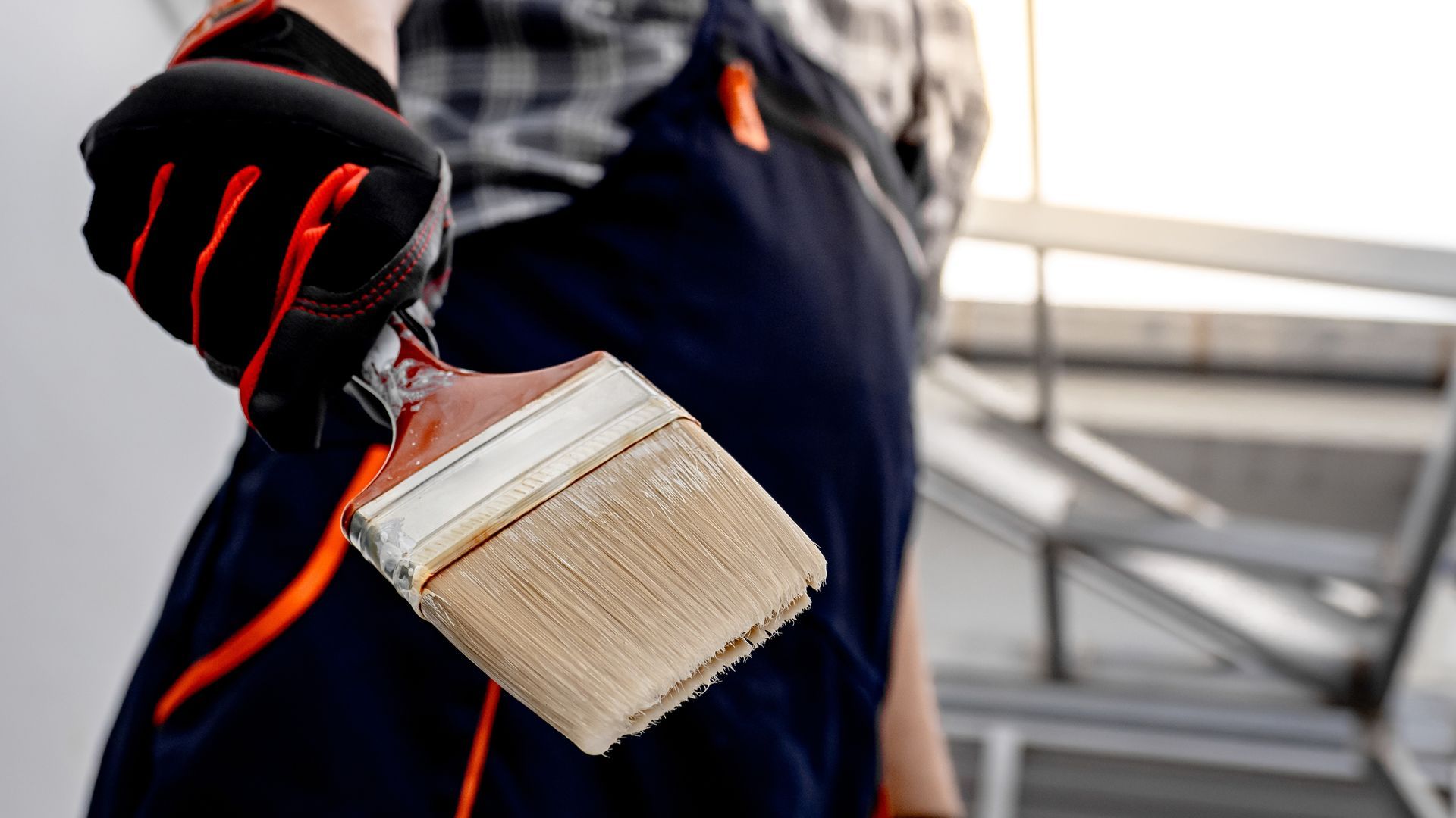 A person wearing work gloves and overalls holds a wide paintbrush against a blurred background.