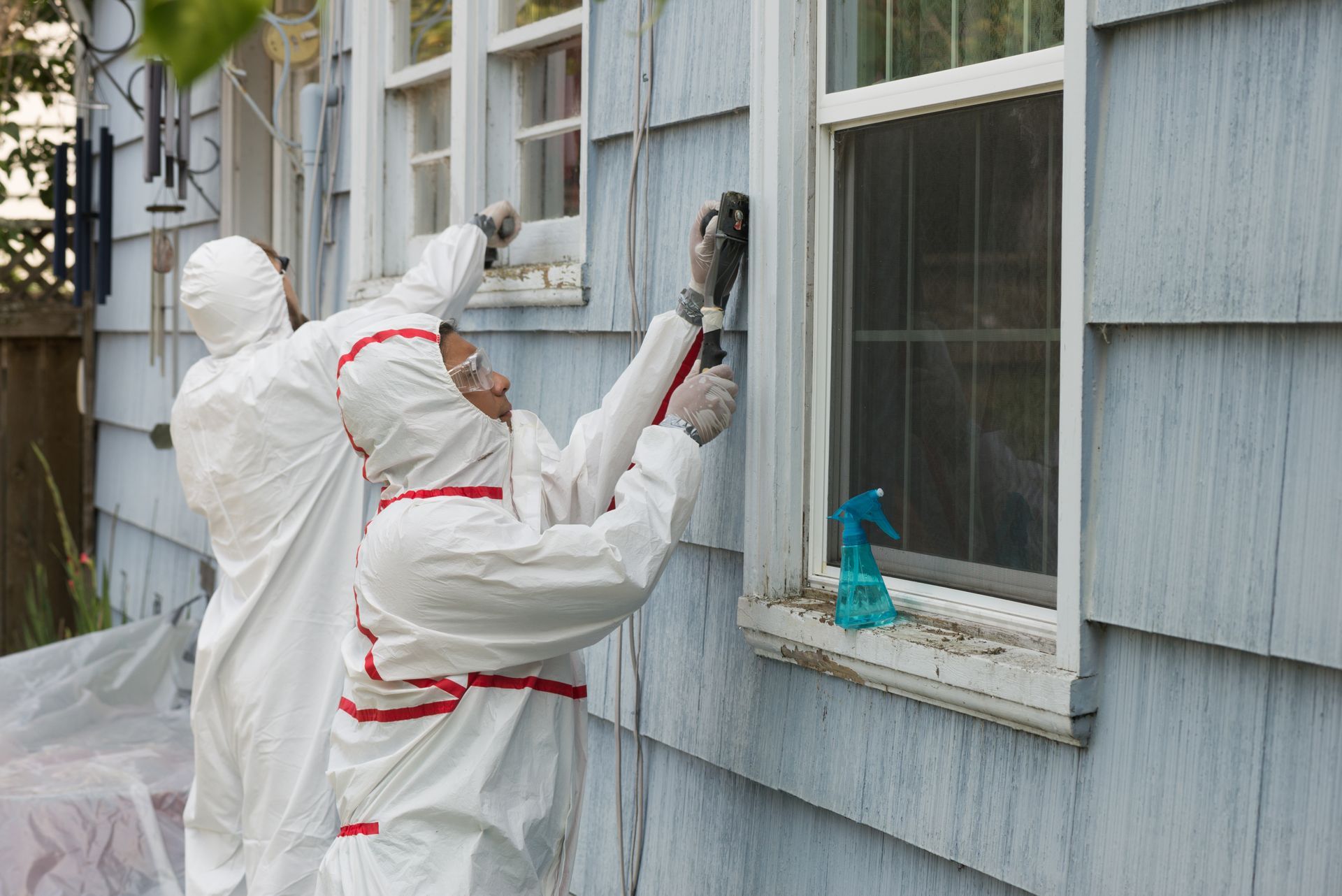 Two workers in full white protective suits and goggles work on the exterior of a light blue house near a window.