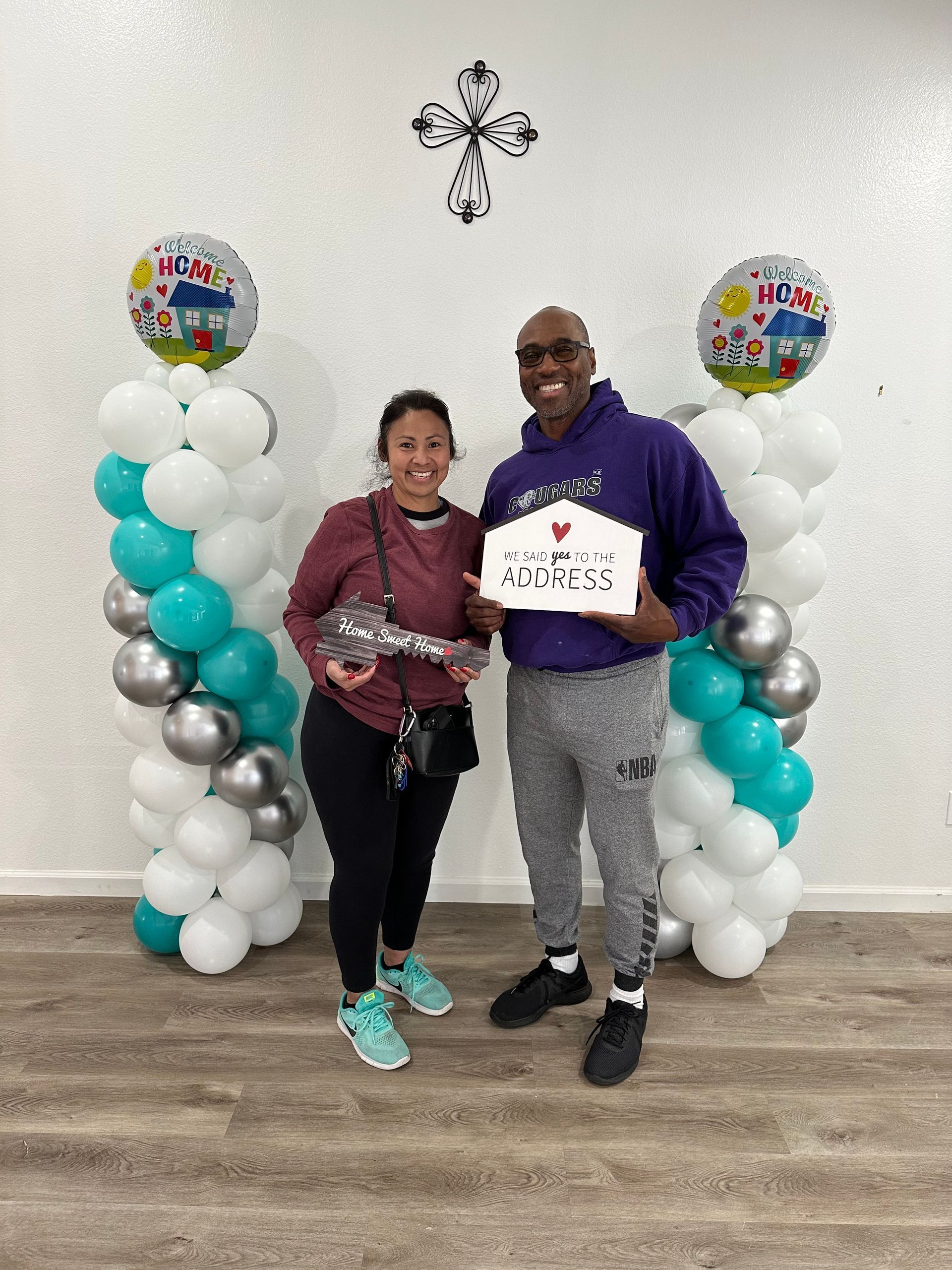Couple holding a house-shaped sign and key, standing by balloon columns indoors, celebrating a new home.