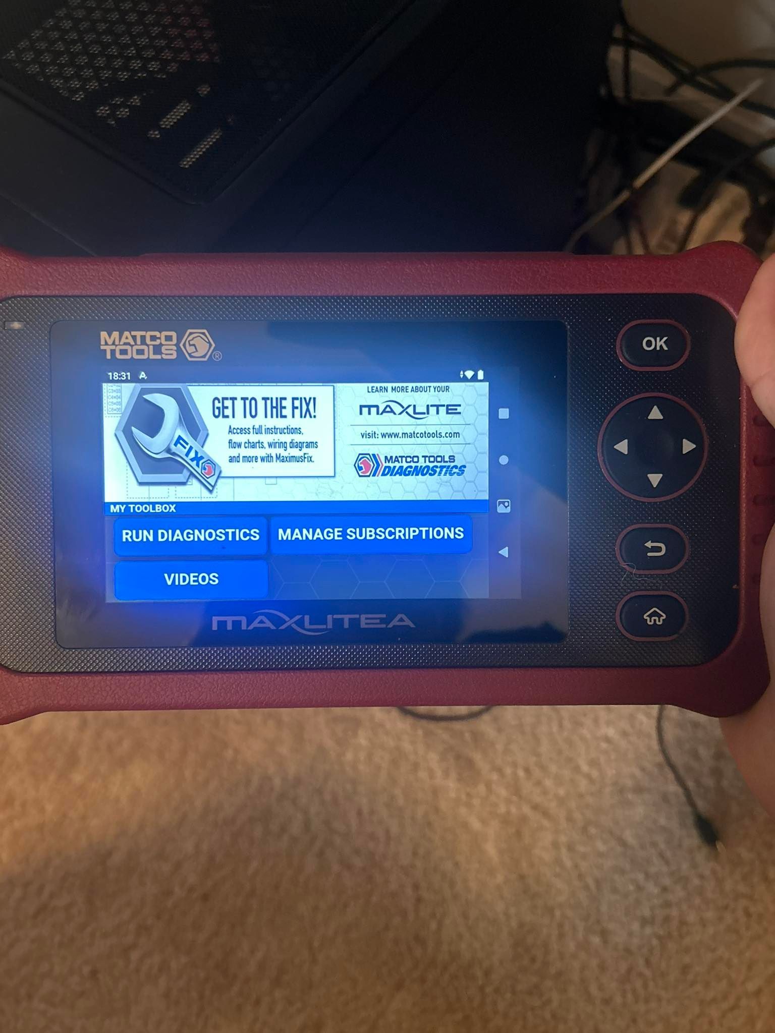 Red and black handheld diagnostic tool showing a menu screen with options. A hand is holding the device.