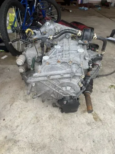 Engine, likely from a vehicle, on a concrete surface with attached hoses and components. A bicycle is in the background.