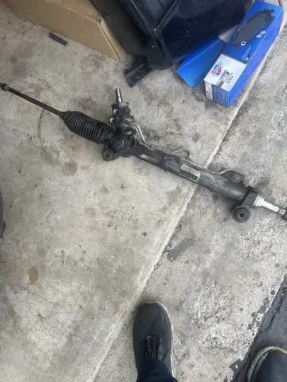 Steering rack on concrete, with black rubber boots and tie rod ends.