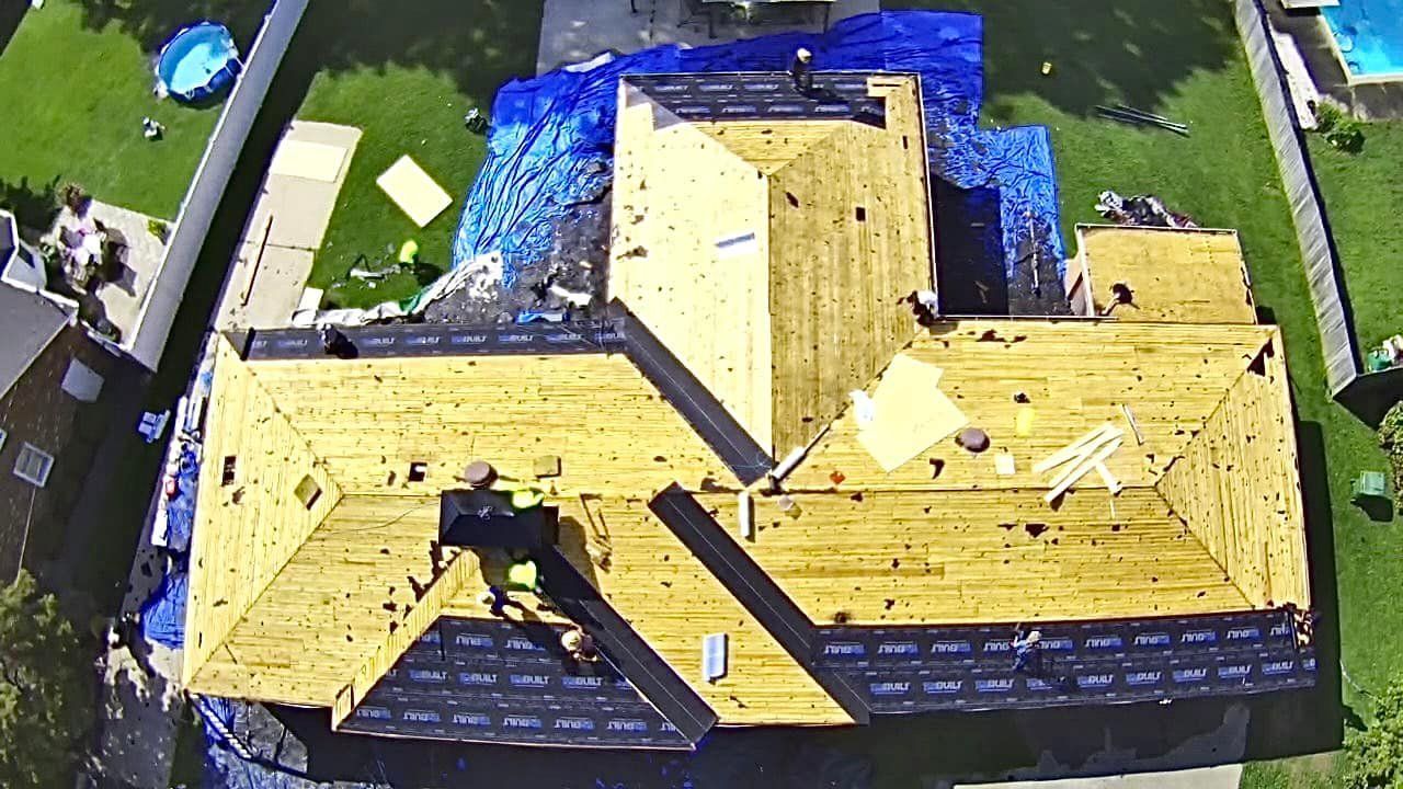 Aerial view of a house with roof construction in progress; workers on roof with blue tarp covering portions.