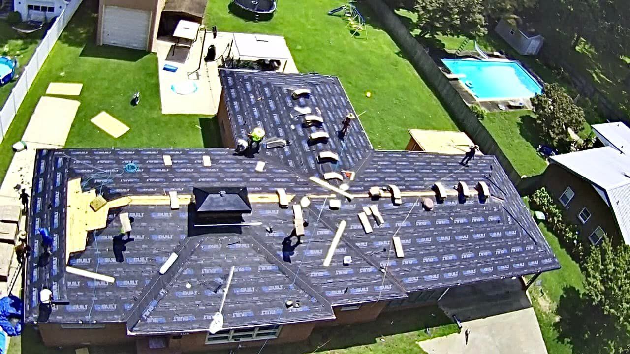 Roofers working on a residential roof, surrounded by lawn. Blue tarp covers parts of the roof.