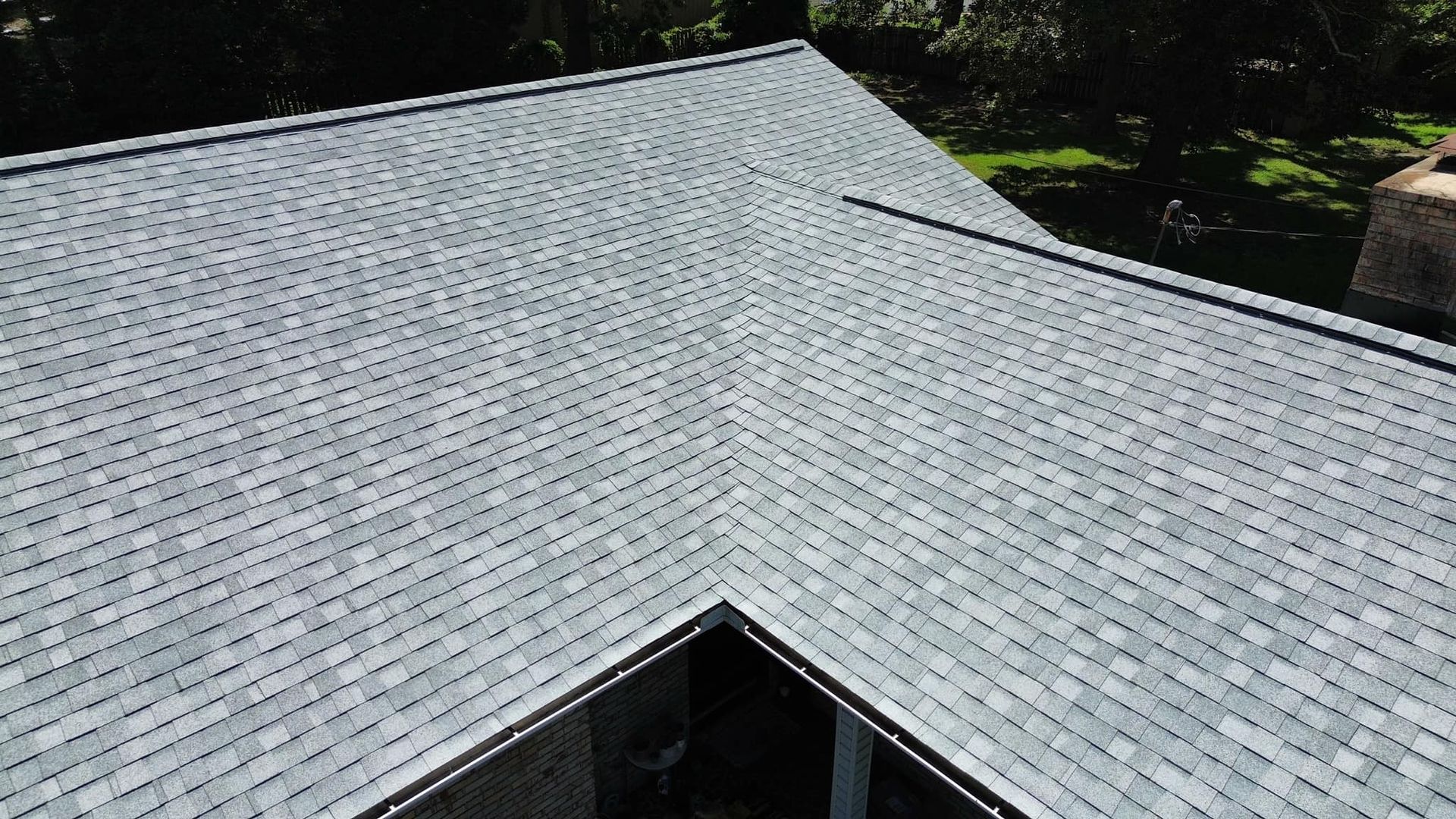 Overhead view of a house roof covered in gray shingles with a triangular shape, surrounded by greenery.