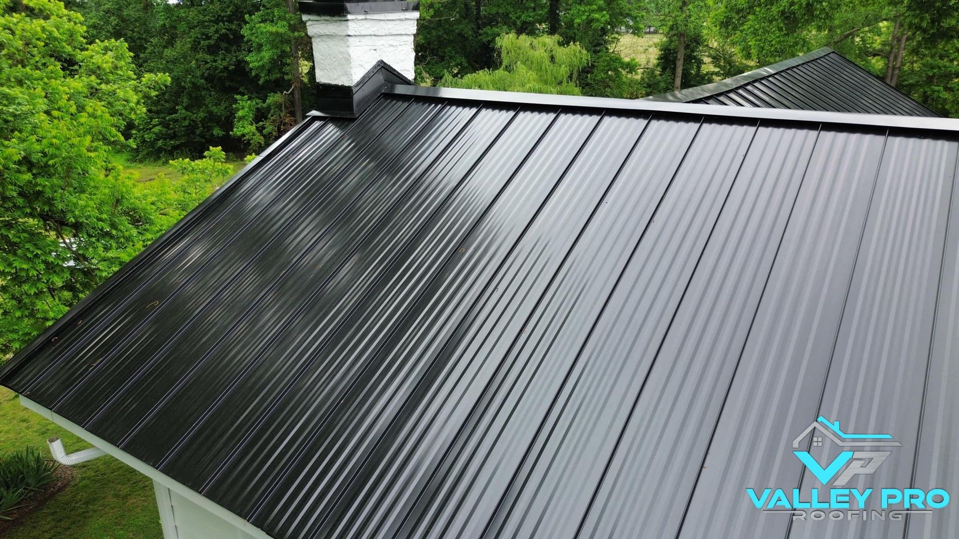 Black metal roof with vertical lines, next to a white chimney and green trees.