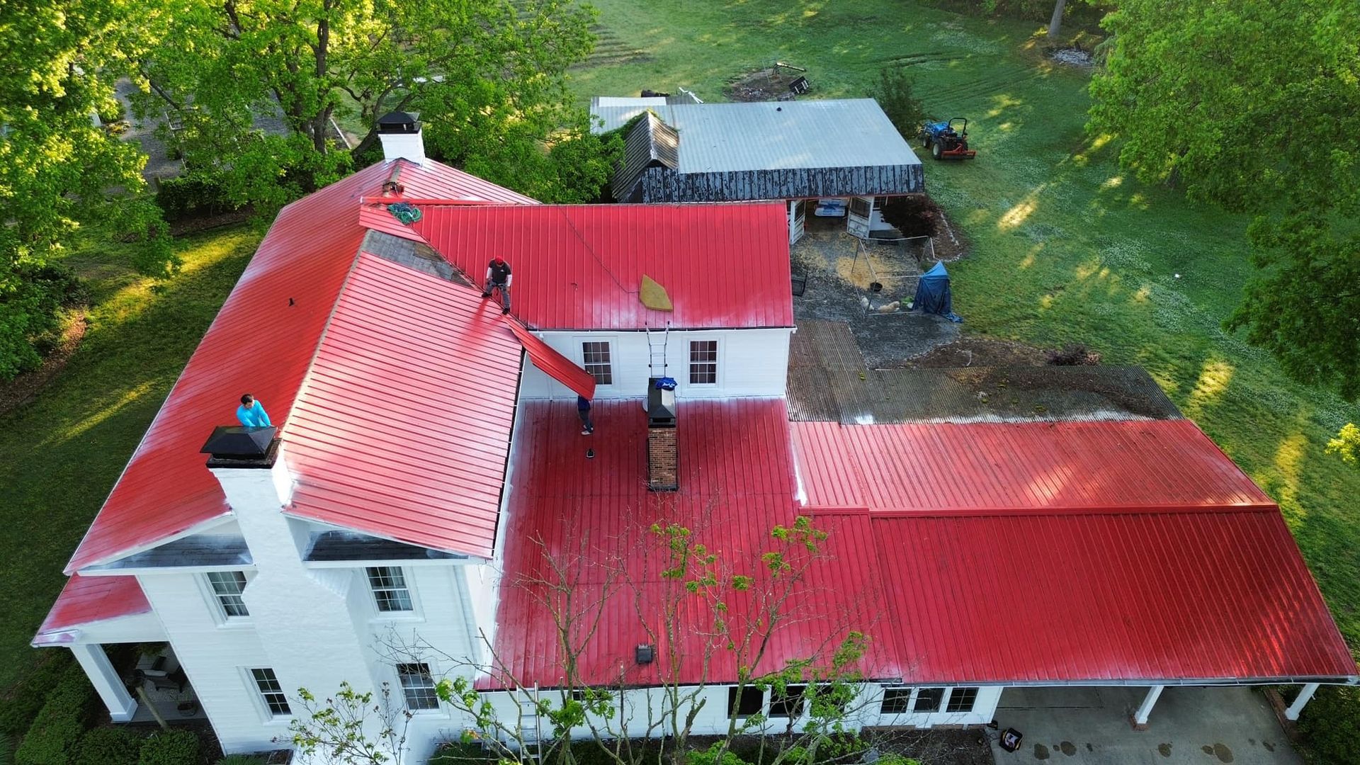 Aerial view of a white house with a red metal roof, surrounded by green trees and grass.
