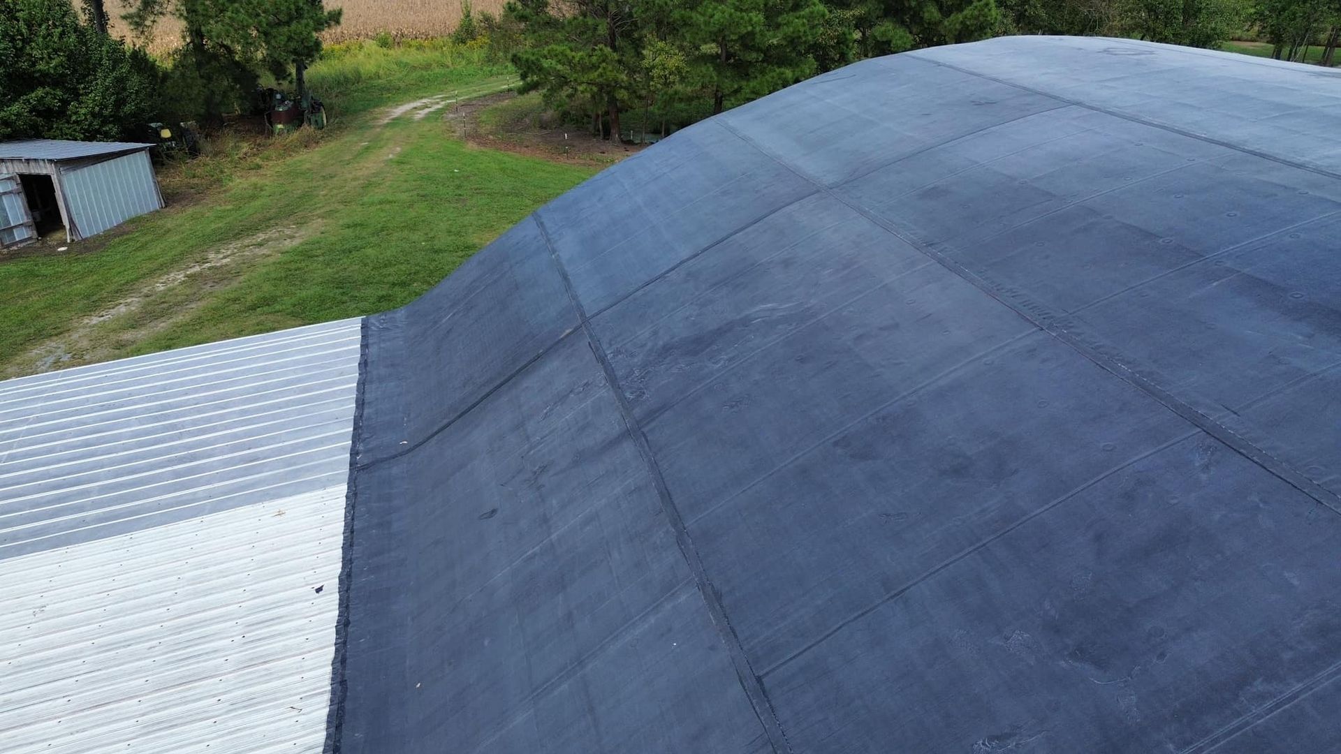 Black, curved roof on a building, with a silver metal roof section visible. Green field and trees in the background.