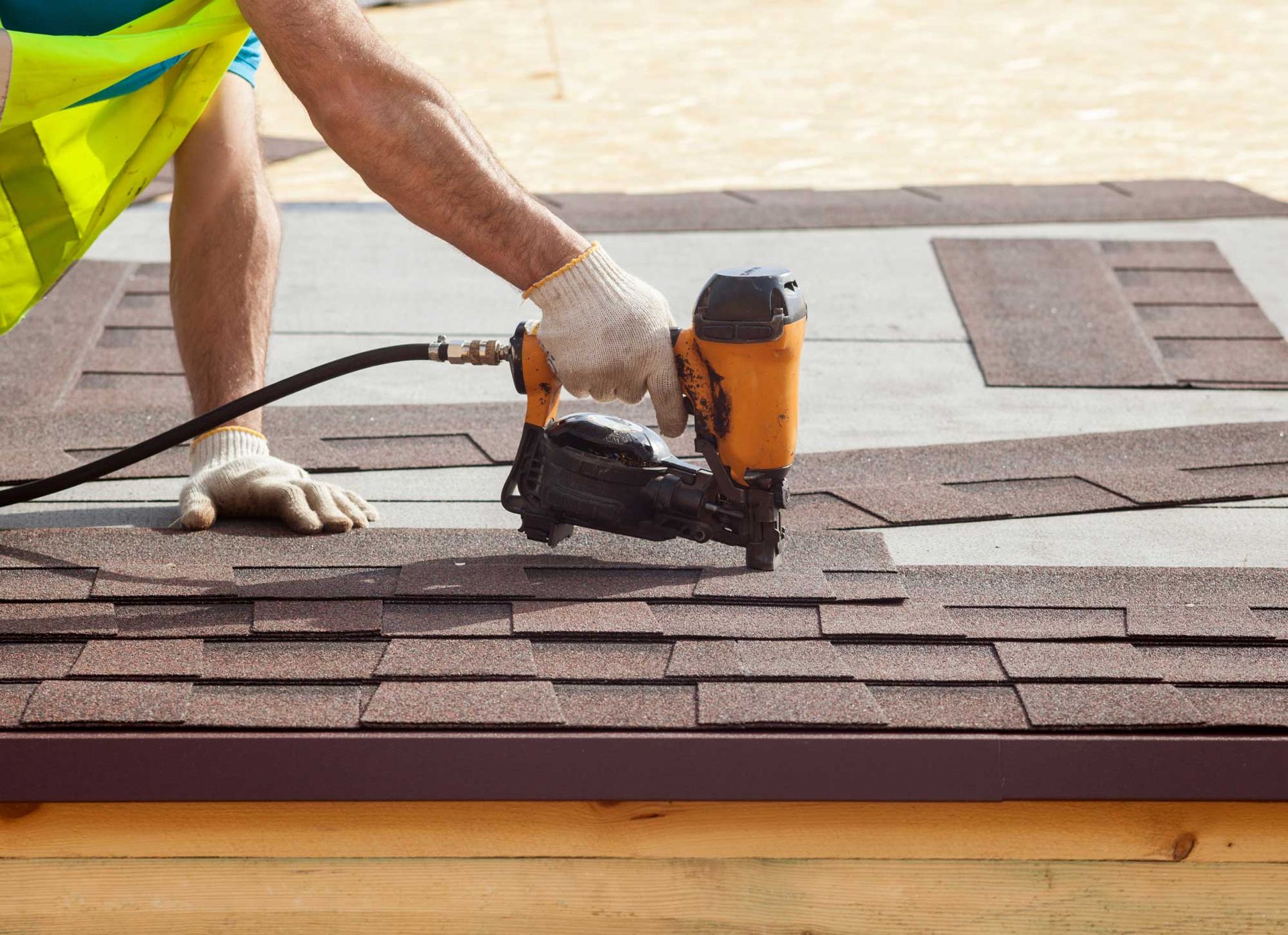A construction worker is putting asphalt roofing shingles with a nail gun.