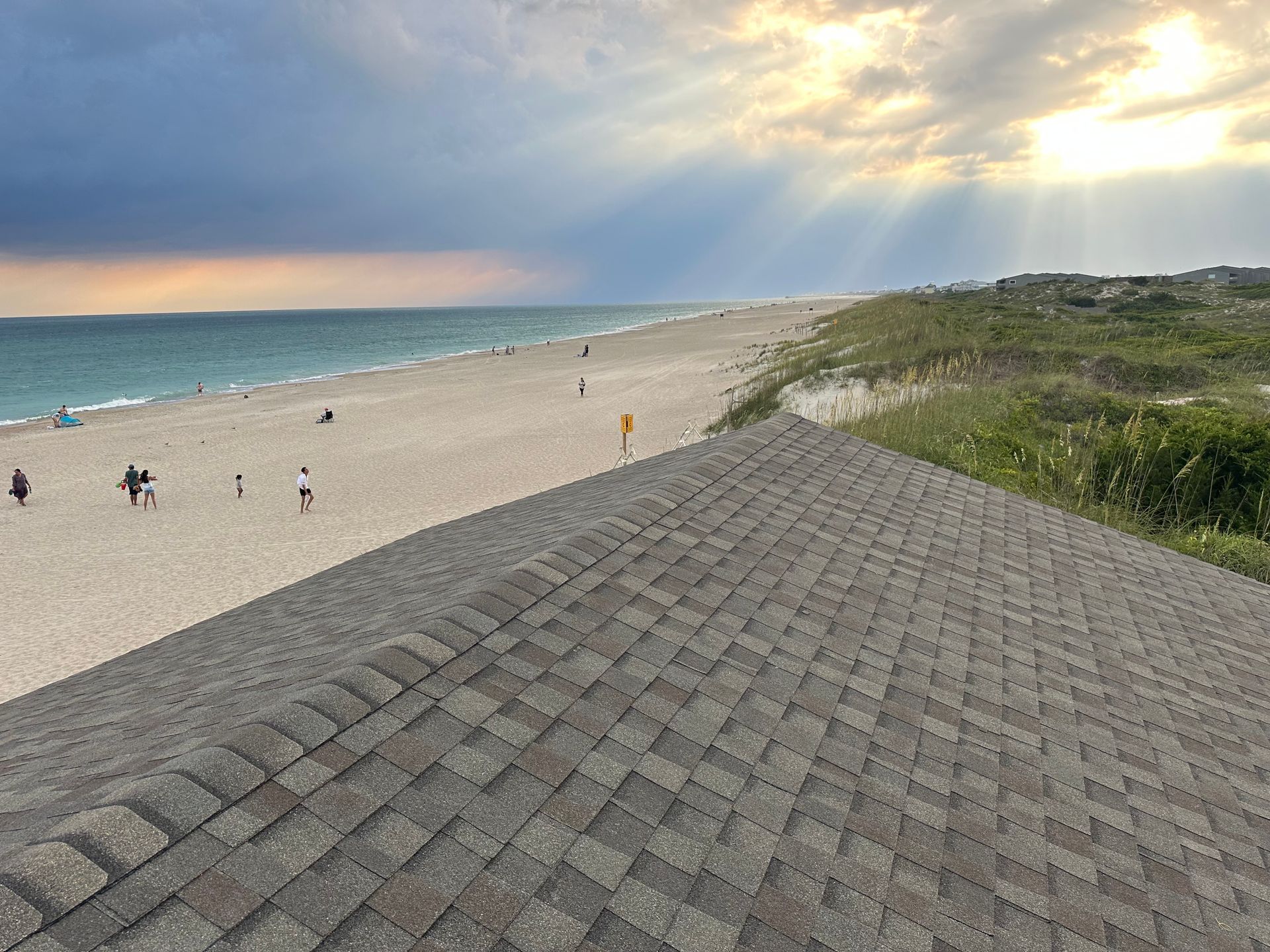 Beach scene: sand, ocean, and sky with sun rays, viewed from a rooftop covered with gray shingles.