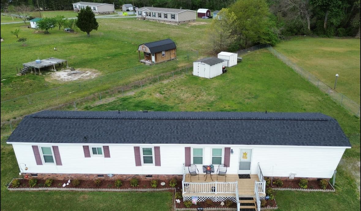 White mobile home with dark roof, small porch, and other buildings on a grassy lot.