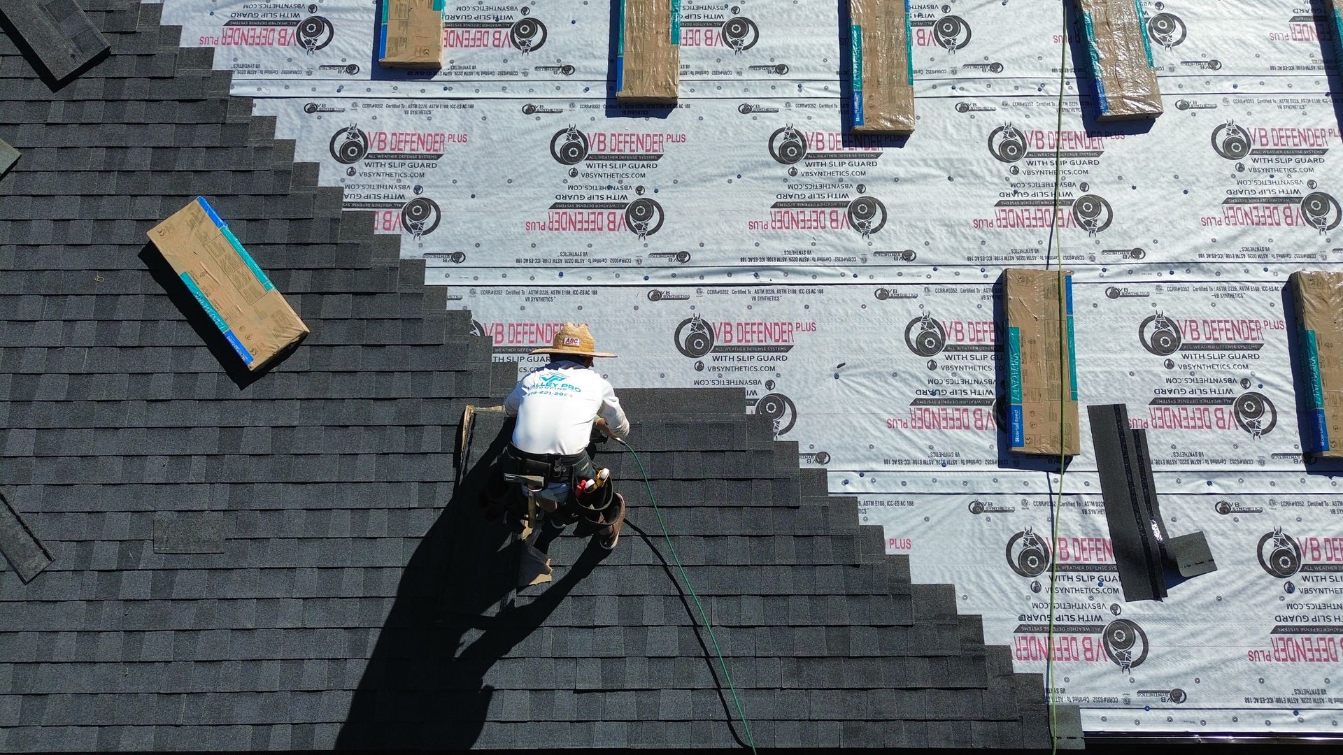 Roofer installing dark gray shingles on a roof, working above a silver underlayment with visible wooden boards.