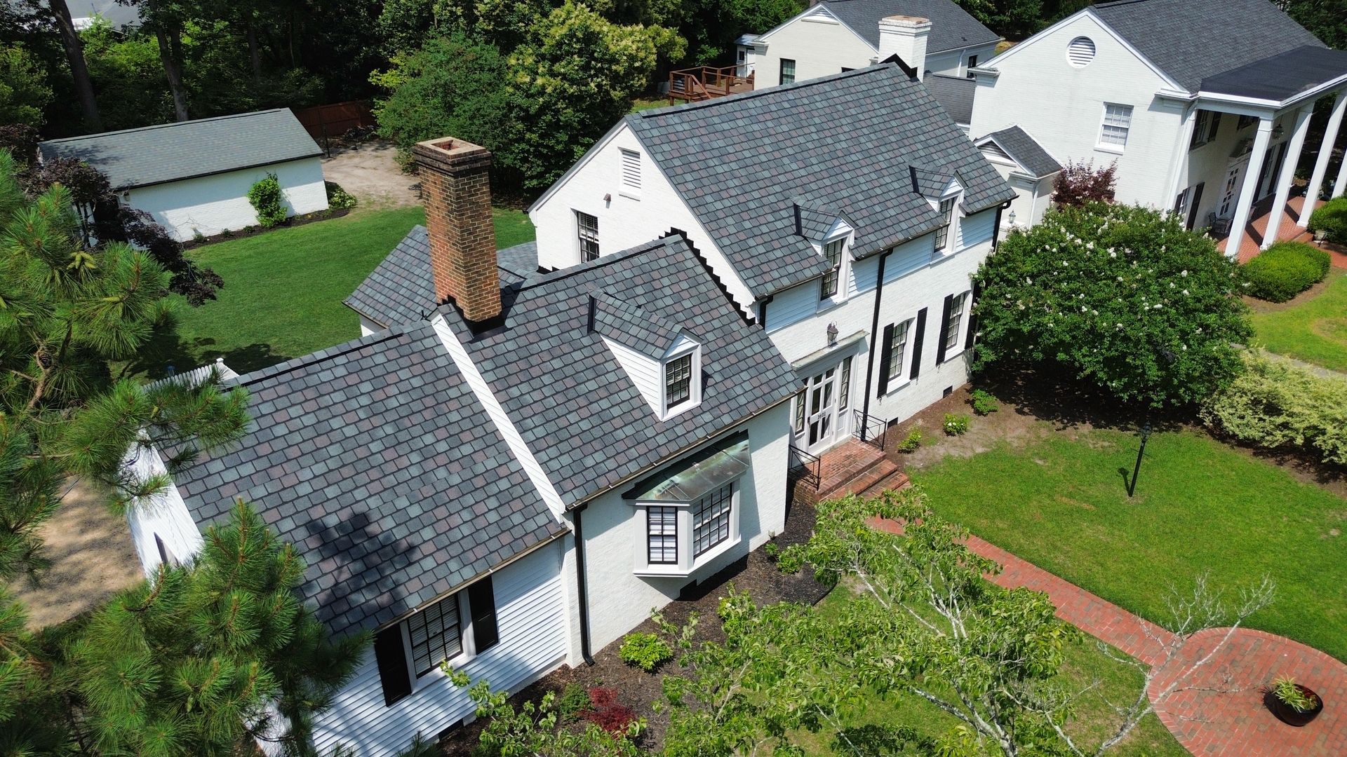 White two-story house with dark gray roof, chimney, and dormers, surrounded by green lawns and trees.