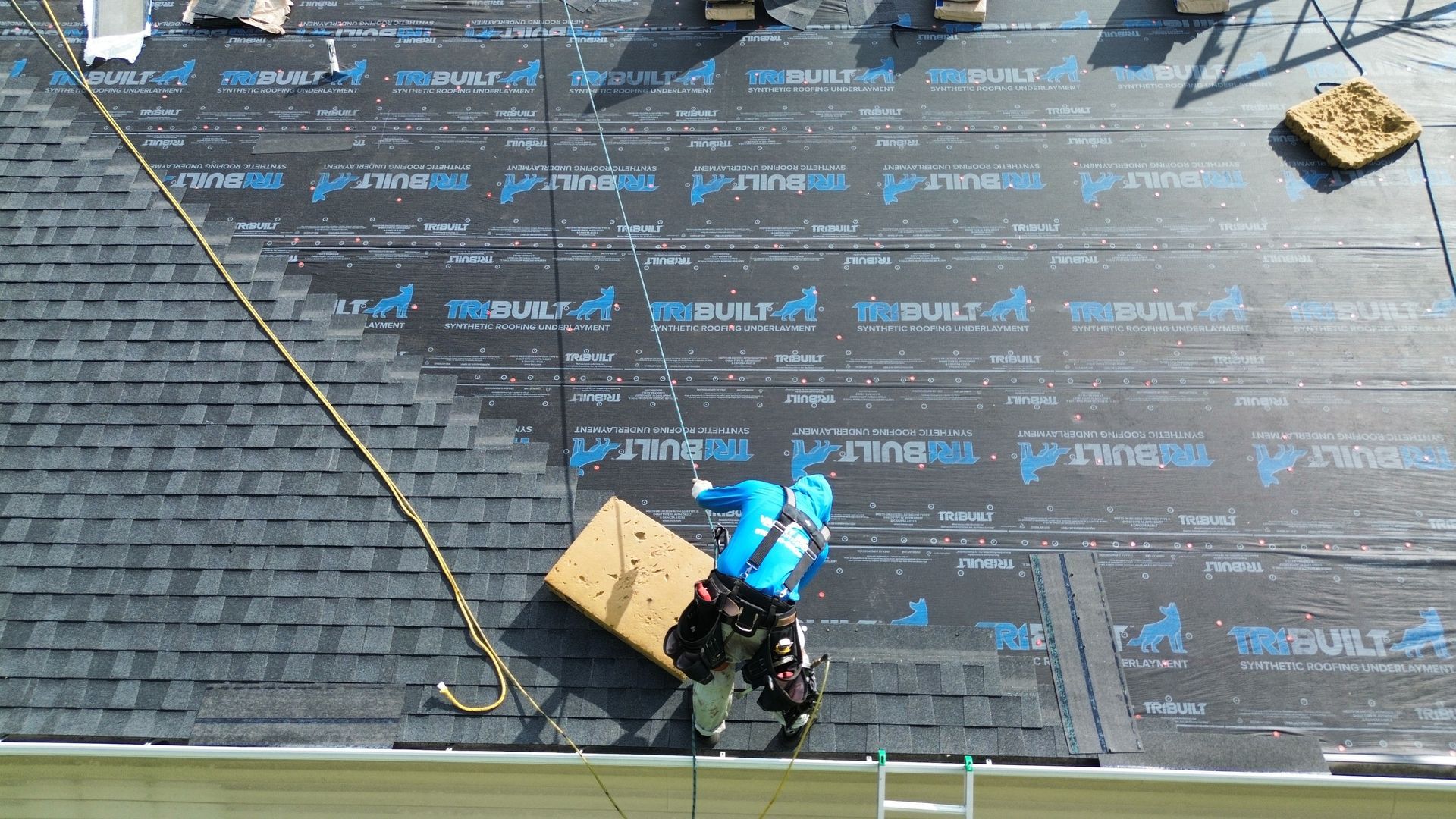 Roofer installing shingles on a roof, wearing safety gear and attached to a safety rope.