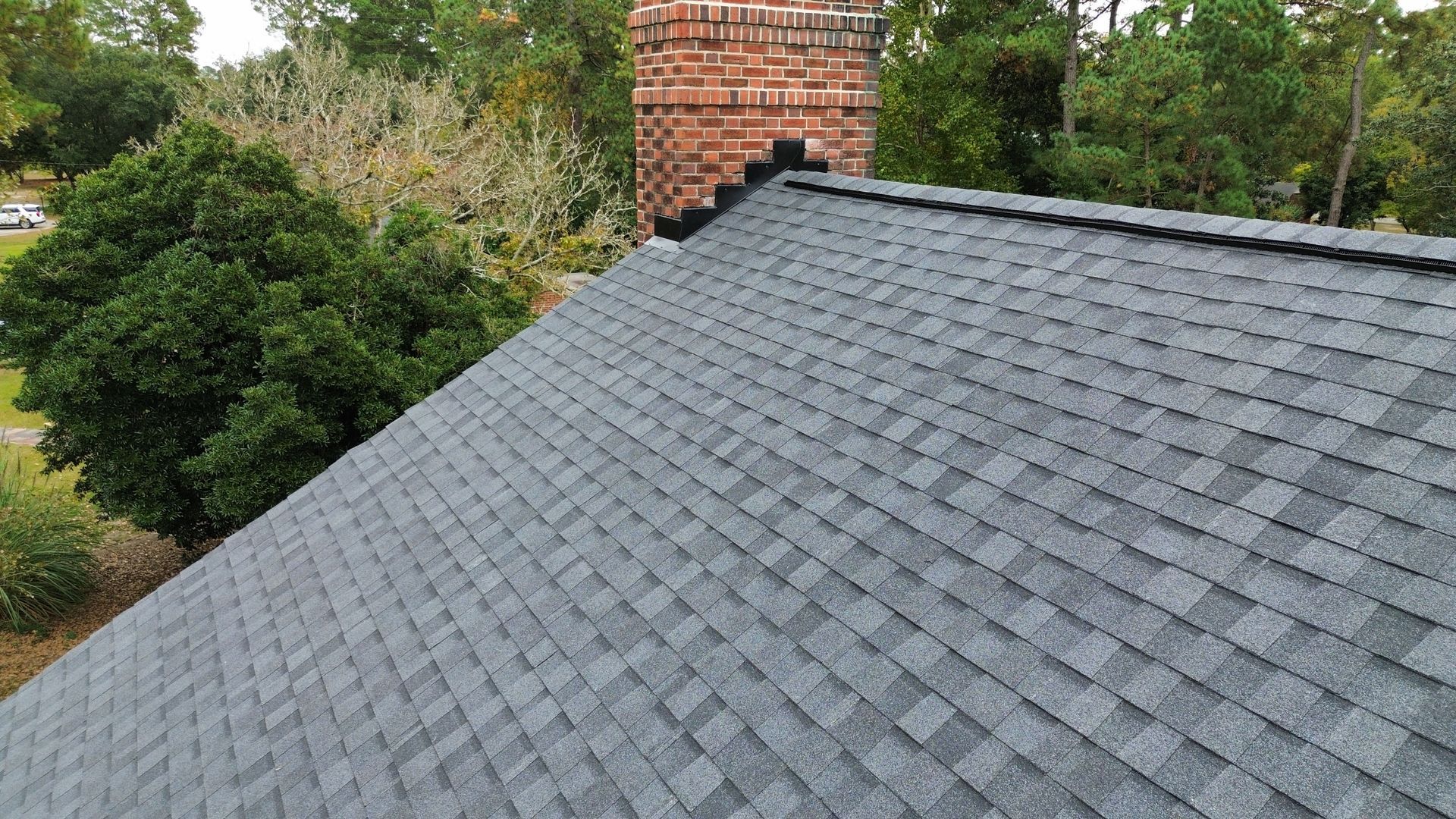 Gray asphalt shingle roof with a brick chimney and green trees in the background.