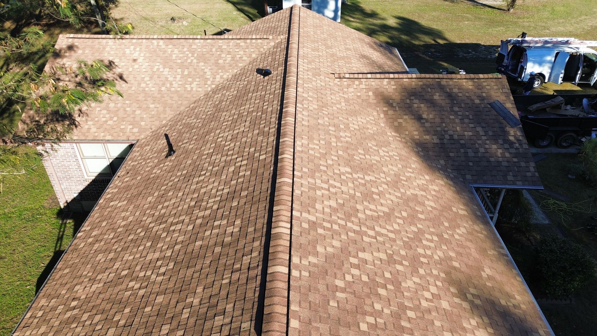 Aerial view of a brown shingle roof on a house, with a central ridge and surrounding greenery.