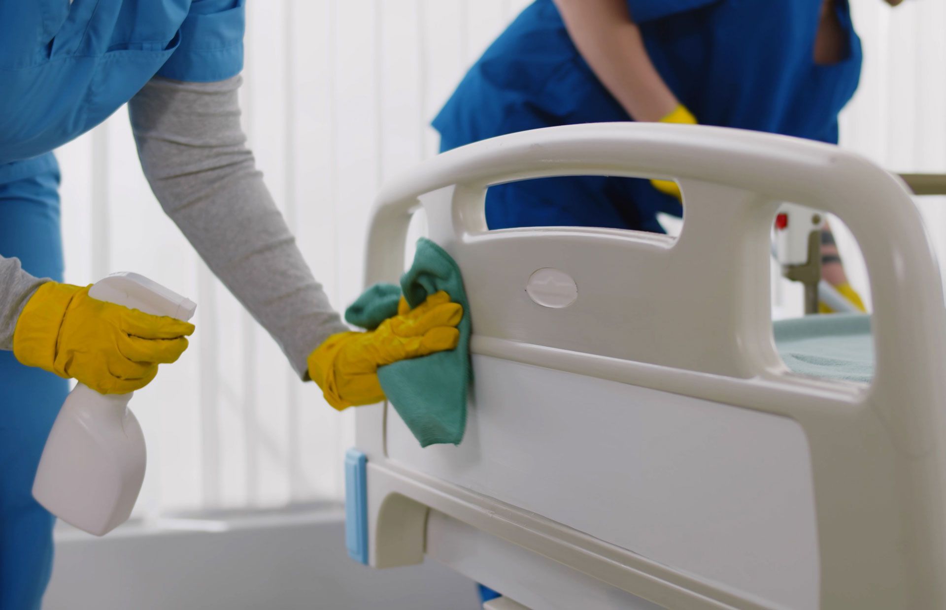 Two people in blue scrubs cleaning a white hospital bed with a spray bottle and cloth.