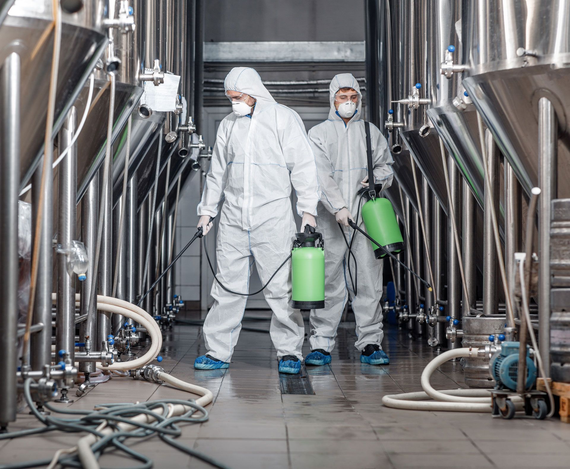 Two workers in protective suits spraying disinfectant in an industrial facility with stainless steel tanks.