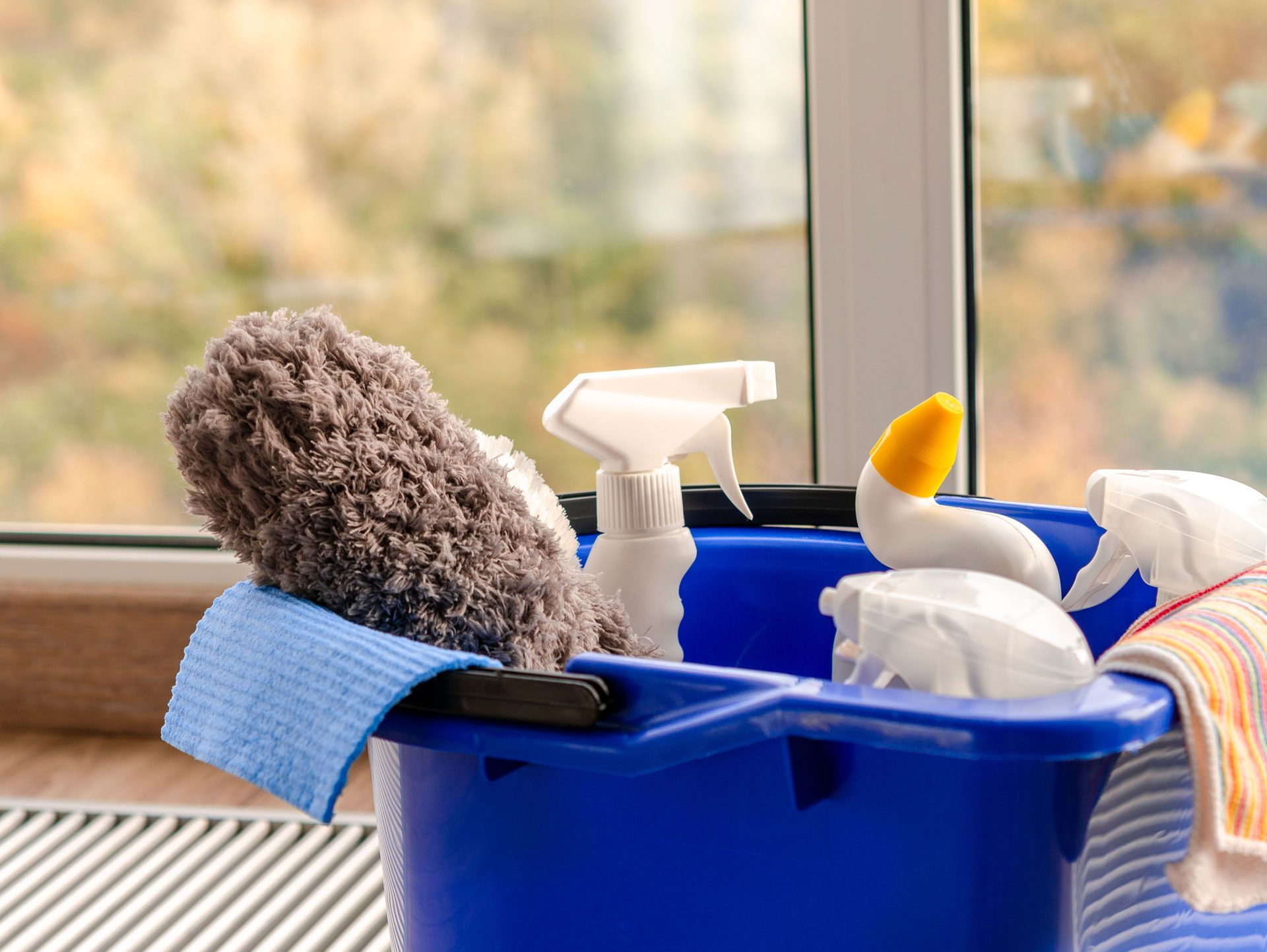 Blue bucket filled with cleaning supplies near a window: microfiber cloth, spray bottles, and a sponge.