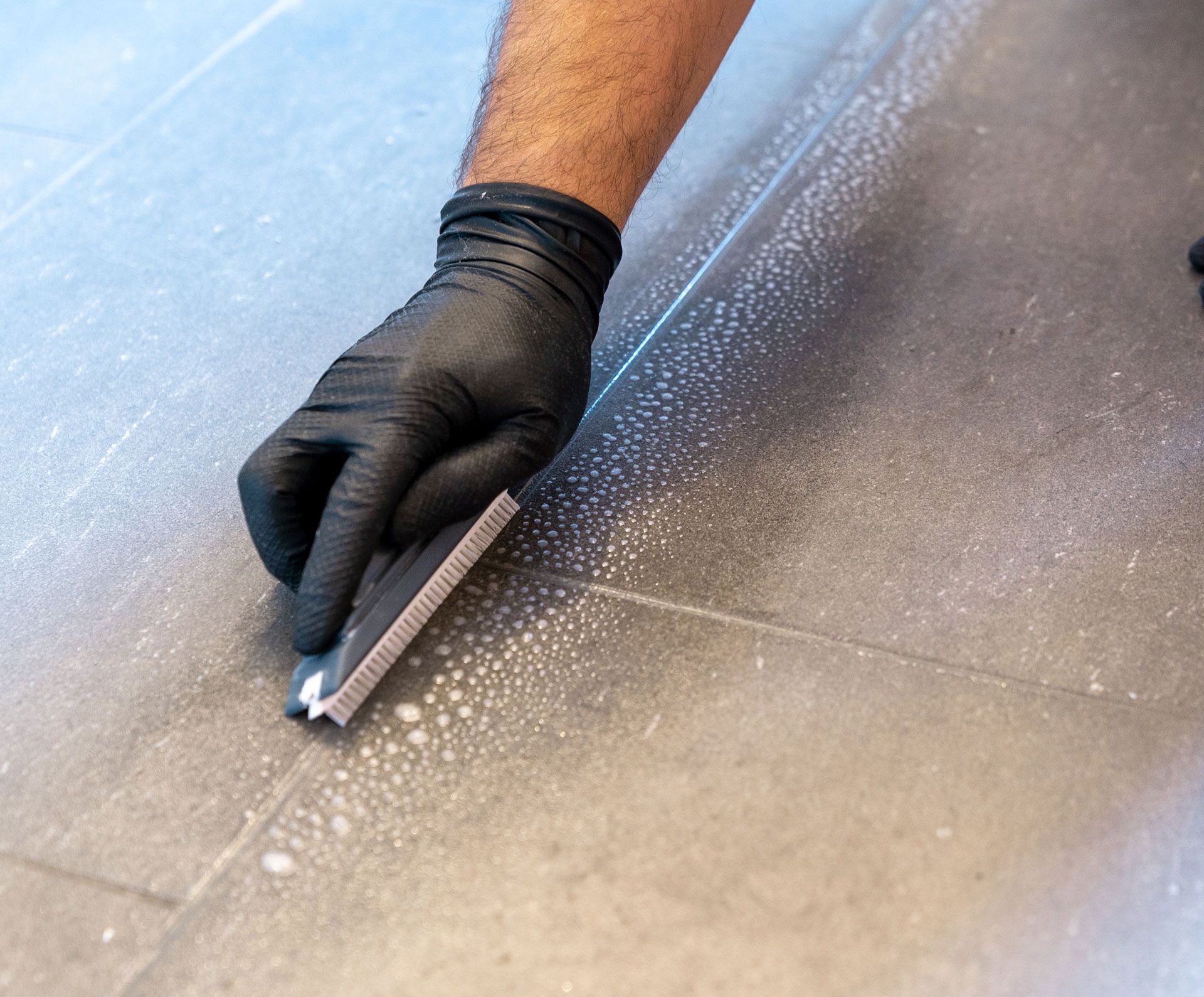 Gloved hand using a tool to clean grout between gray floor tiles.
