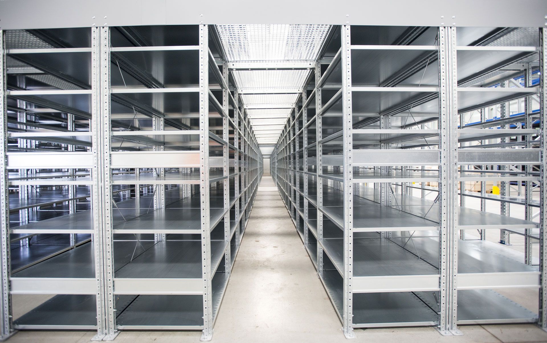 Rows of empty metal shelves in a warehouse aisle, concrete floor, overhead lighting.