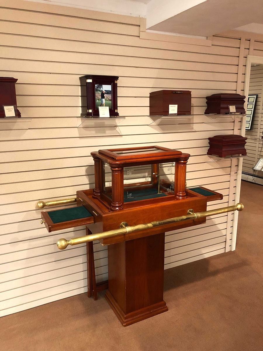 Wooden cremation urns and displays in a showroom. A central podium holds a large ornate urn, with shelves of smaller urns on the wall.