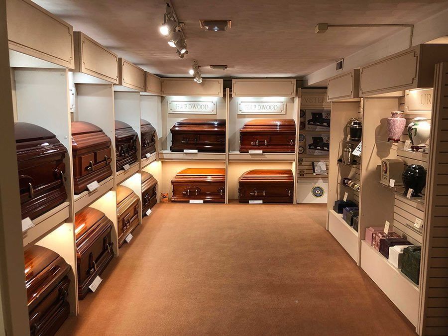 Funeral home display room with coffins, urns, and shelves. Brown and beige tones, carpeted floor.