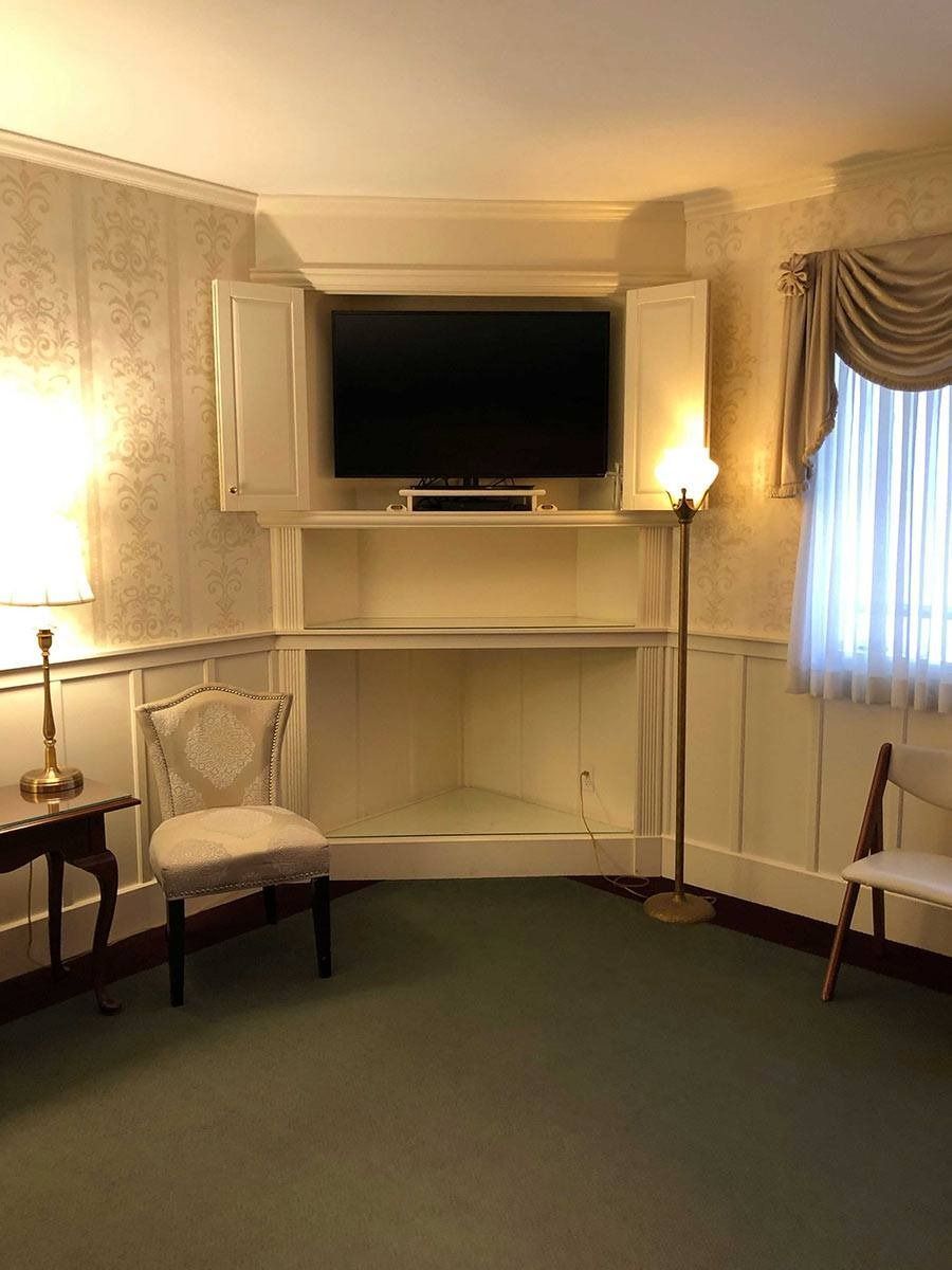 Corner room with TV in cabinet, chairs, lamp, window with curtain, wallpaper, and green carpet.