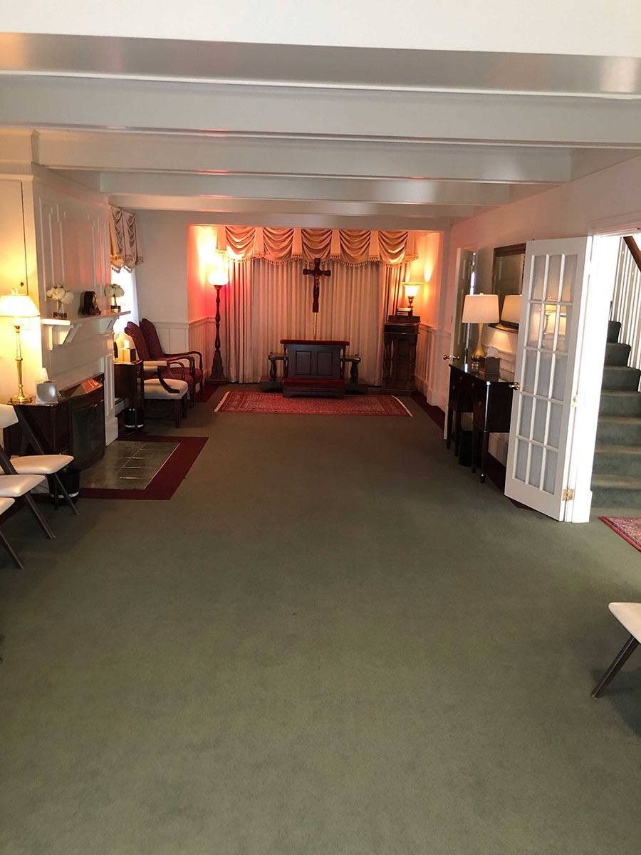 Interior of a chapel with a cross, altar, chairs, and stairs, with green carpet and warm lighting.