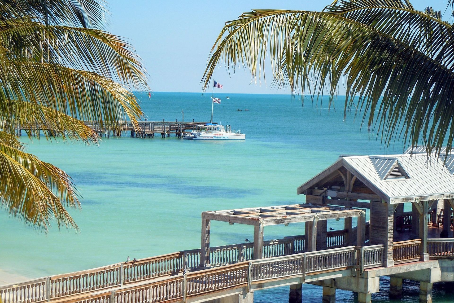 A photo taken through palm trees in the foreground of a couple piers in crystal blue water with a boat in the background.