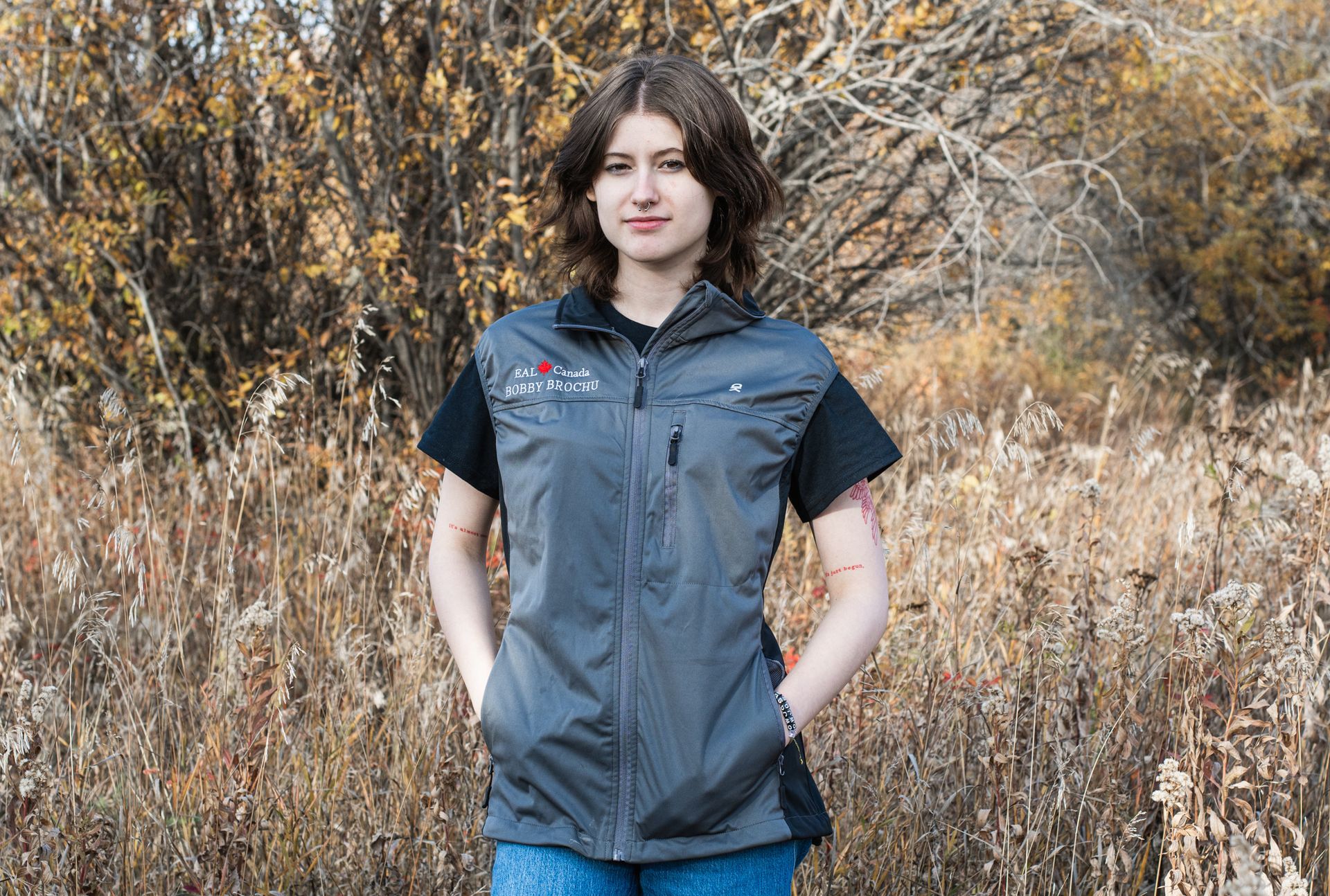 A woman wearing a gray vest is standing in a field with her hands in her pockets.