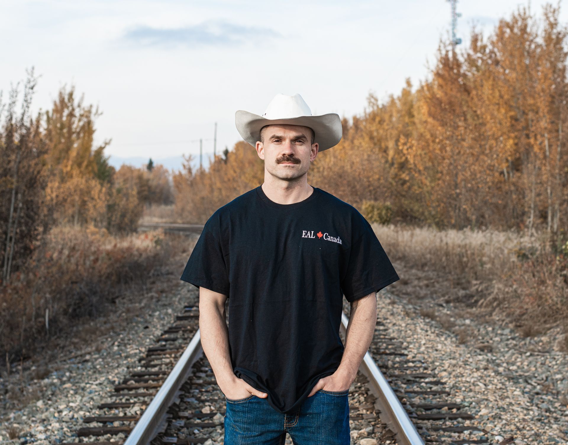 A man wearing a cowboy hat and a black shirt is standing on train tracks.