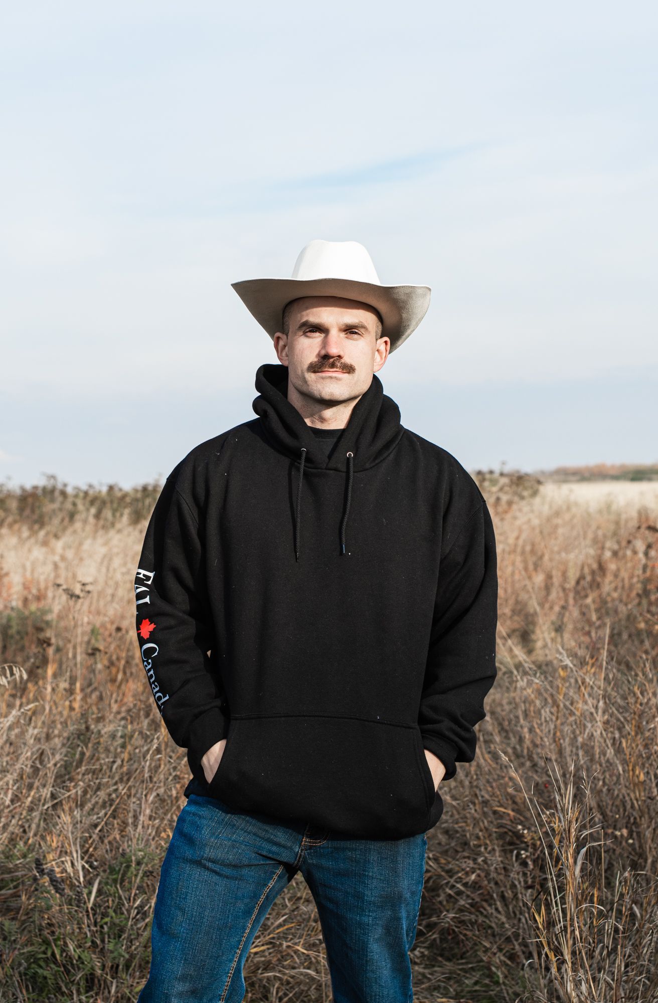 A man wearing a cowboy hat and a black hoodie is standing in a field.