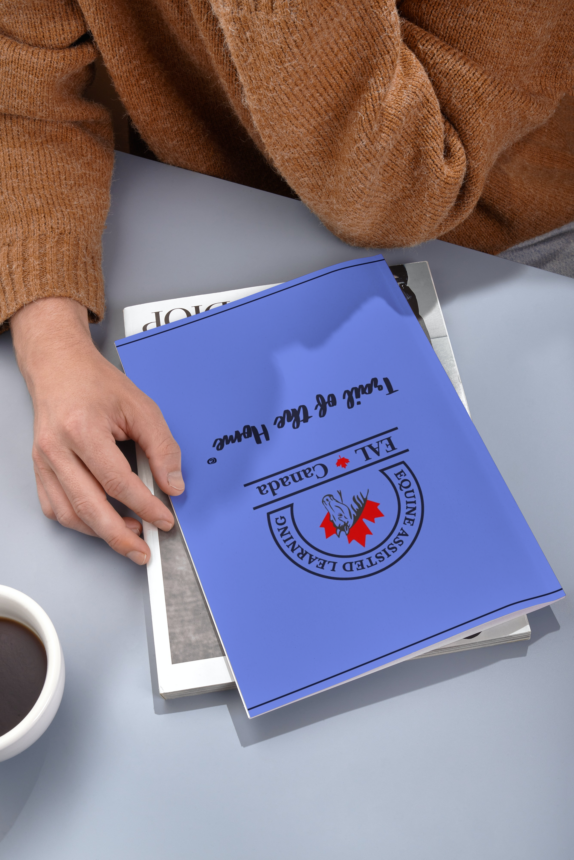 A person is holding a blue book on a table next to a cup of coffee.