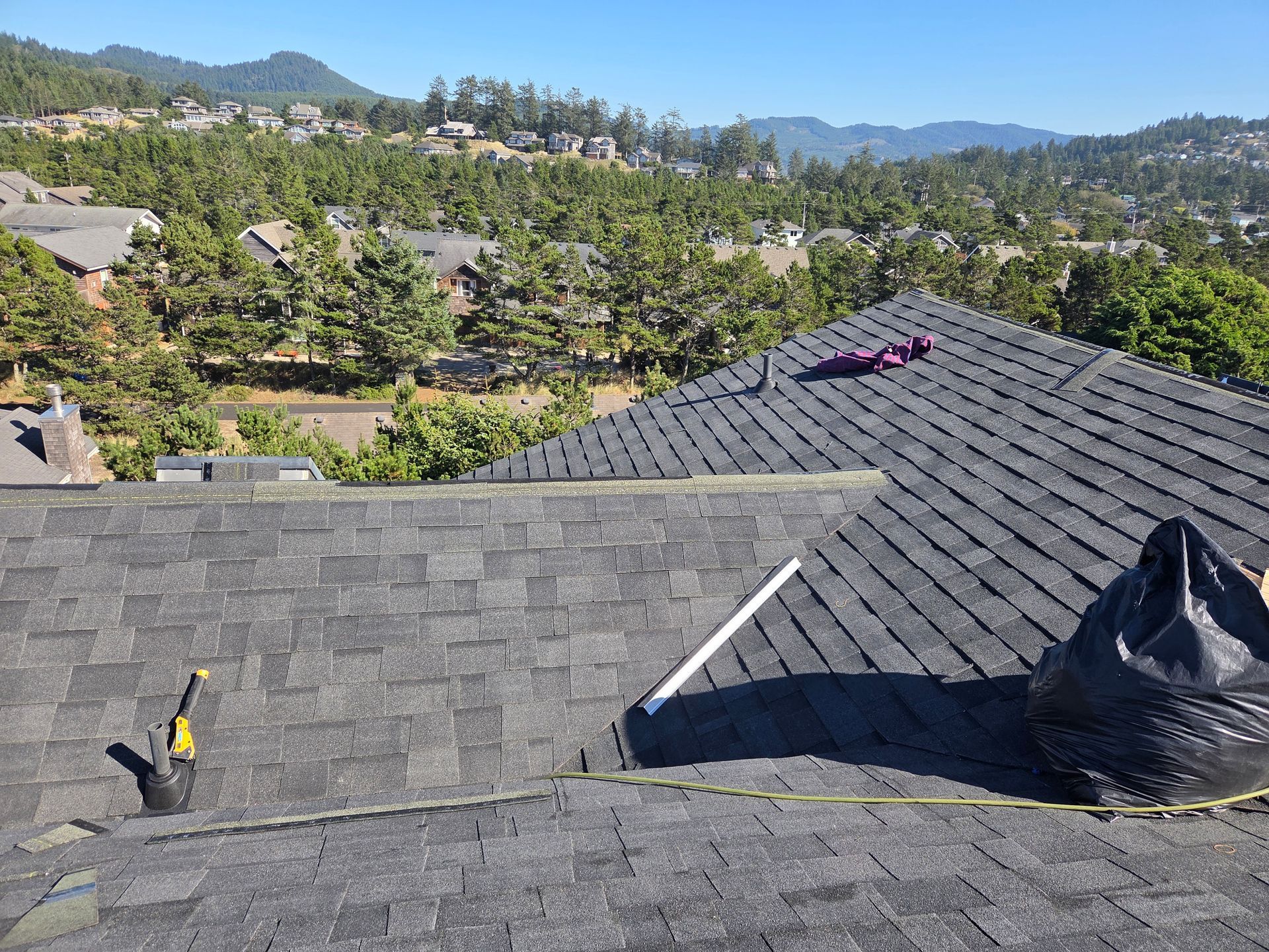 Rooftop with black shingles, tools, and trash bag, overlooking a neighborhood and hills under a blue sky.