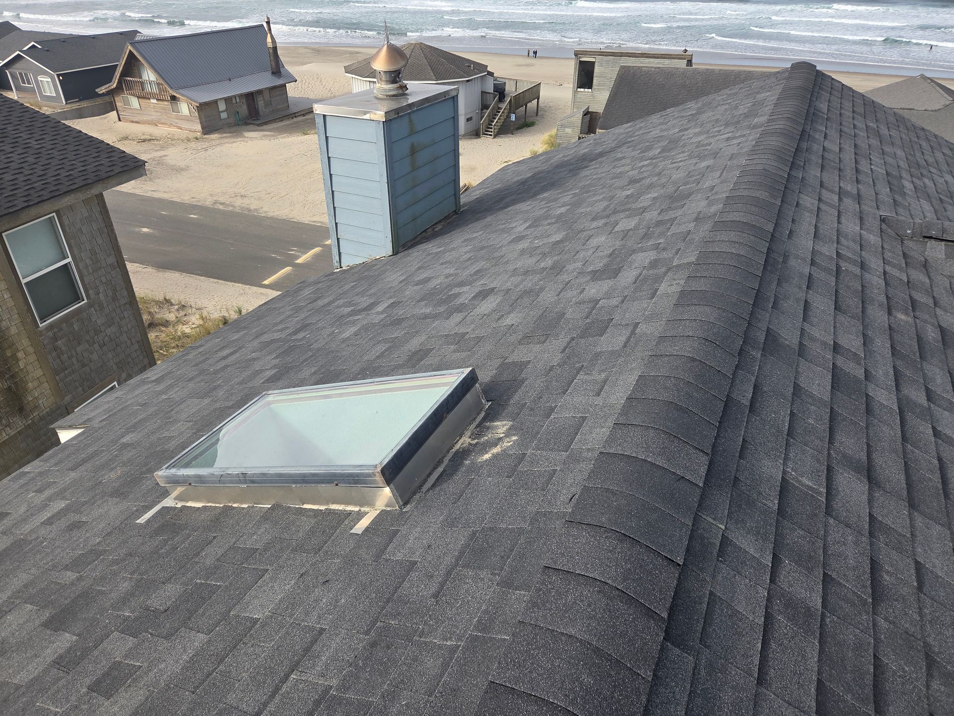 Gray shingle roof with a skylight and chimney, overlooking a sandy beach with ocean and buildings.