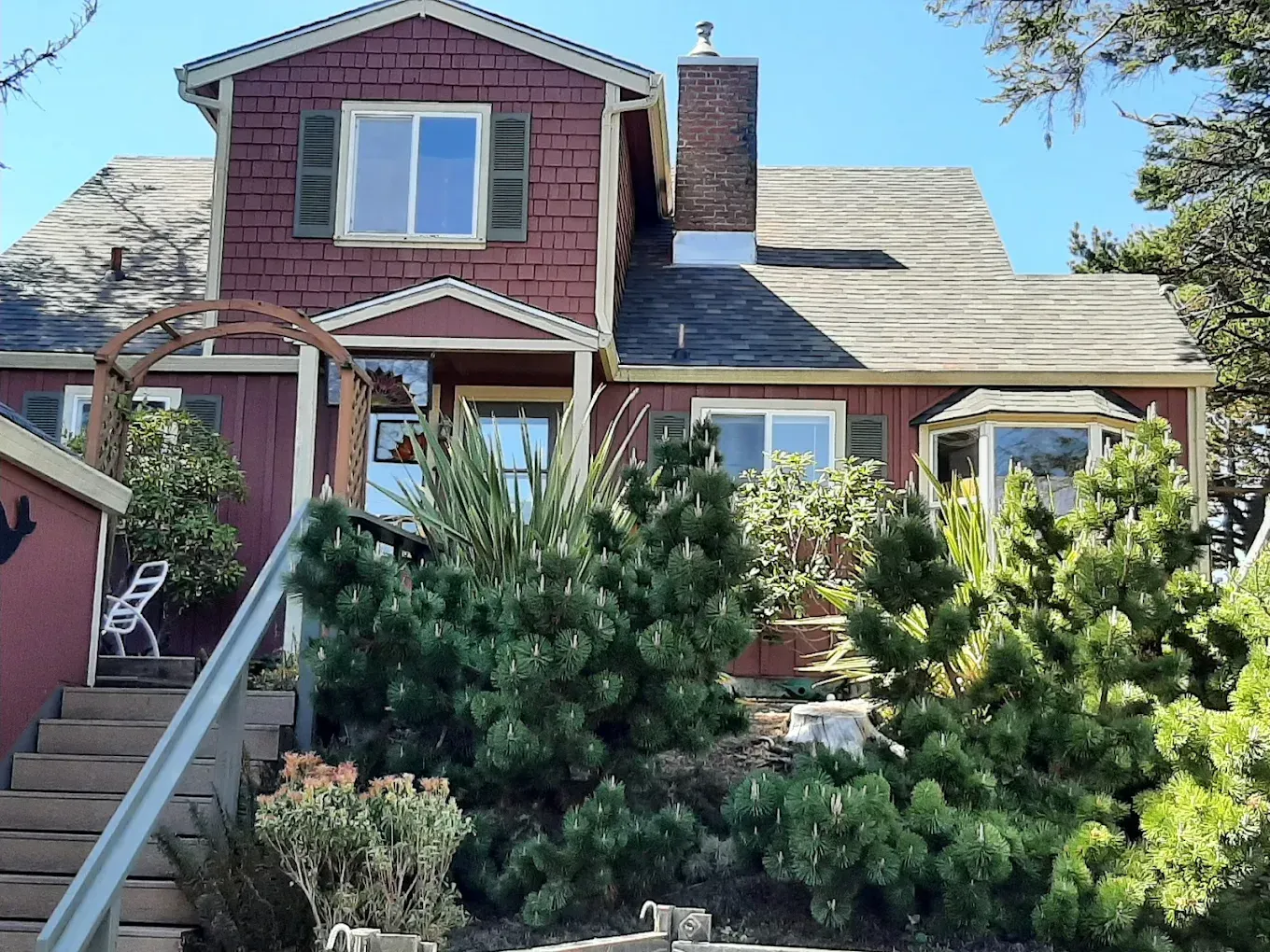 Two-story reddish house with dark shutters, surrounded by evergreen bushes, on a sunny day.
