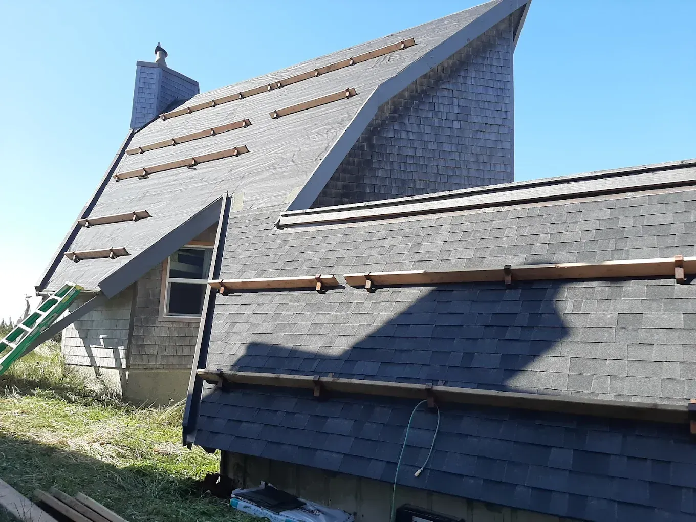 House roof construction; grey shingles, wooden rails, green ladder against the building, clear blue sky.