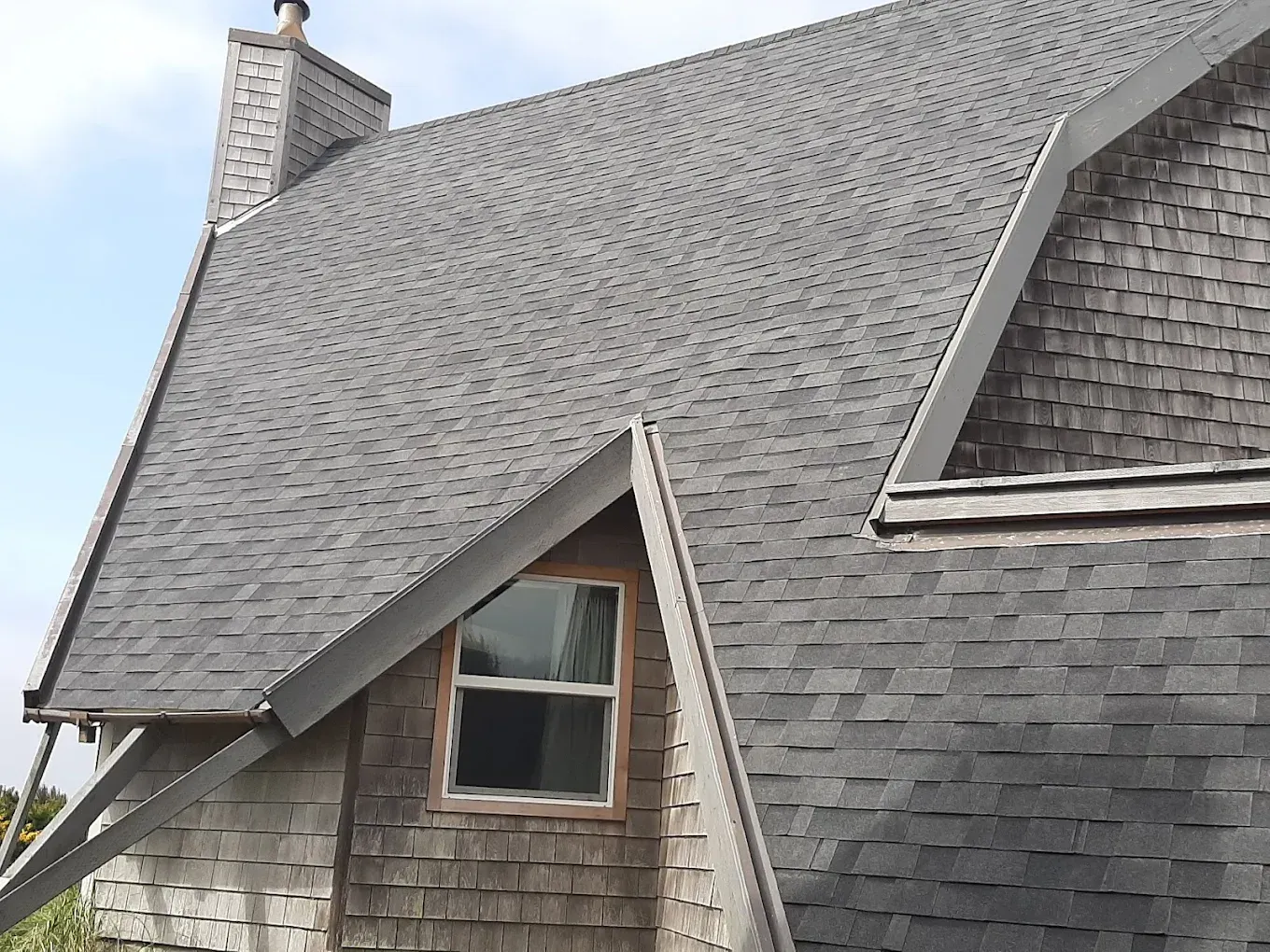 Gray shingled roof with a chimney and a window, on a cloudy day.