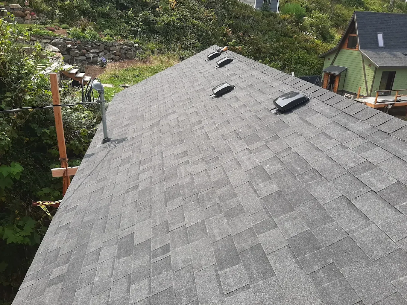 Dark gray shingle roof with vents; a partially built house and trees in the background.